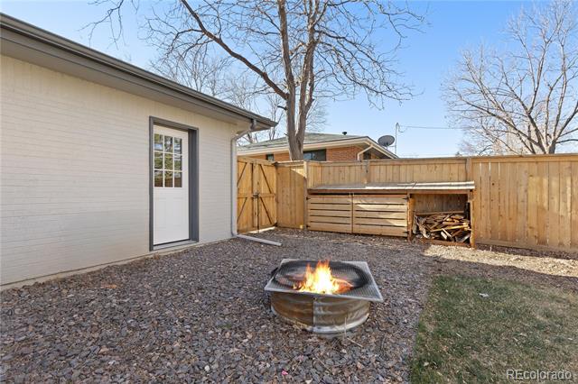 Backyard fire pit burning on a gravel patio beside a wooden fence and wood storage.