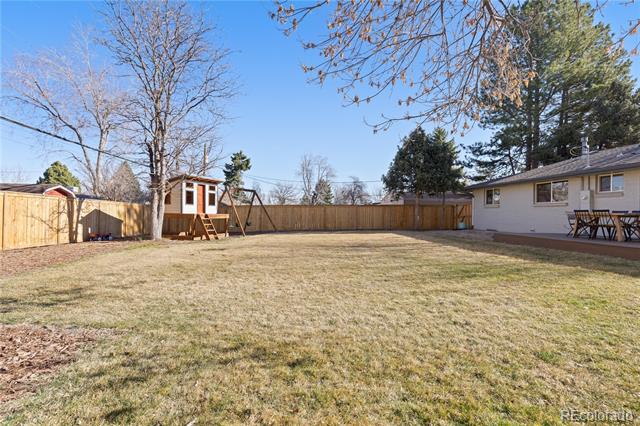 Fenced backyard with a broad lawn, playhouse, swing set, and patio dining area.