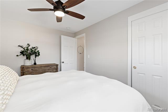 Bedroom with a large white bed, rustic dresser, potted plant, and dark-bladed ceiling fan.