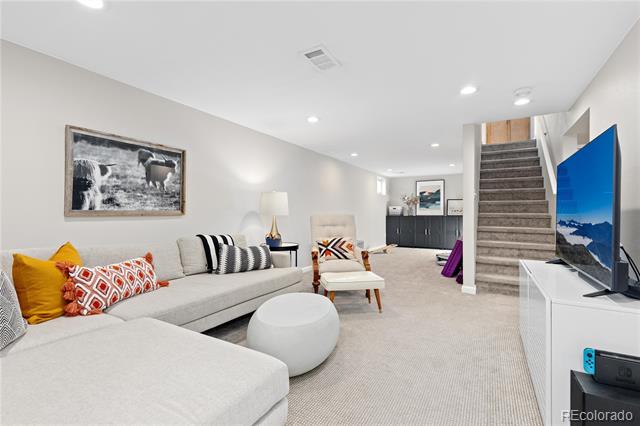 Basement living room with a light sectional, round ottoman, television, and carpeted staircase.