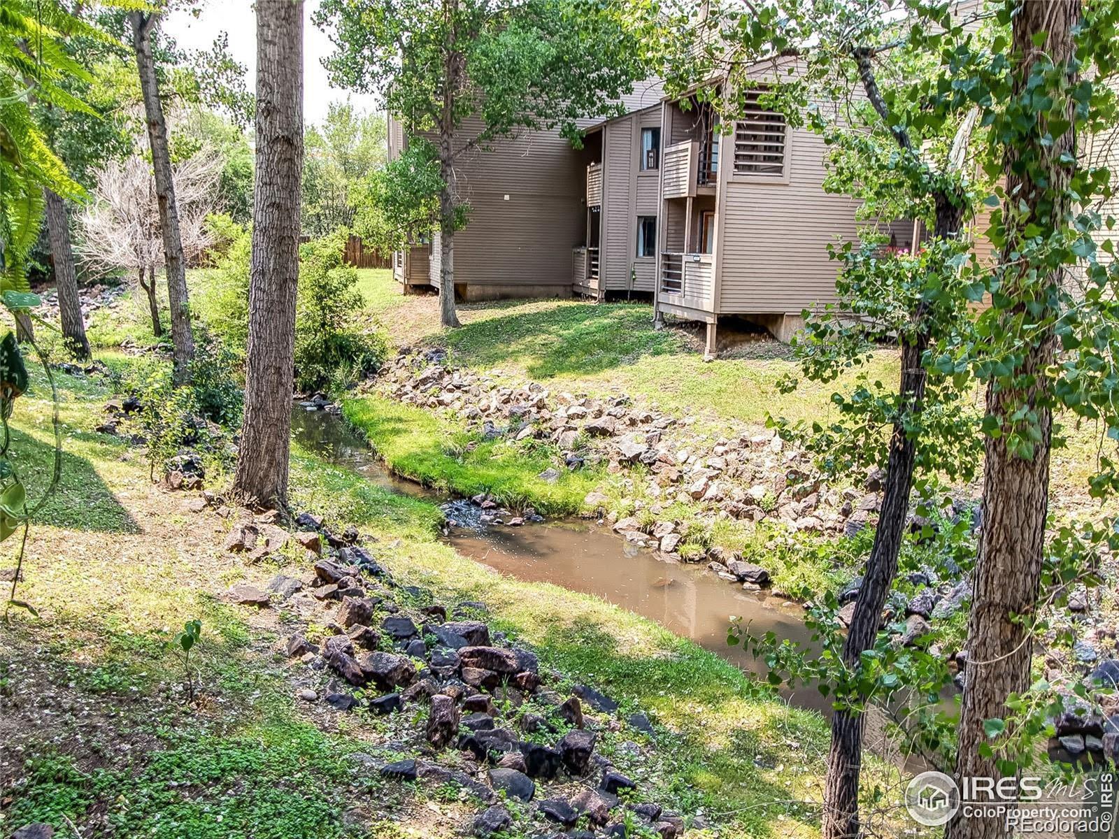 Condominium buildings beside a rock-lined creek shaded by mature trees.