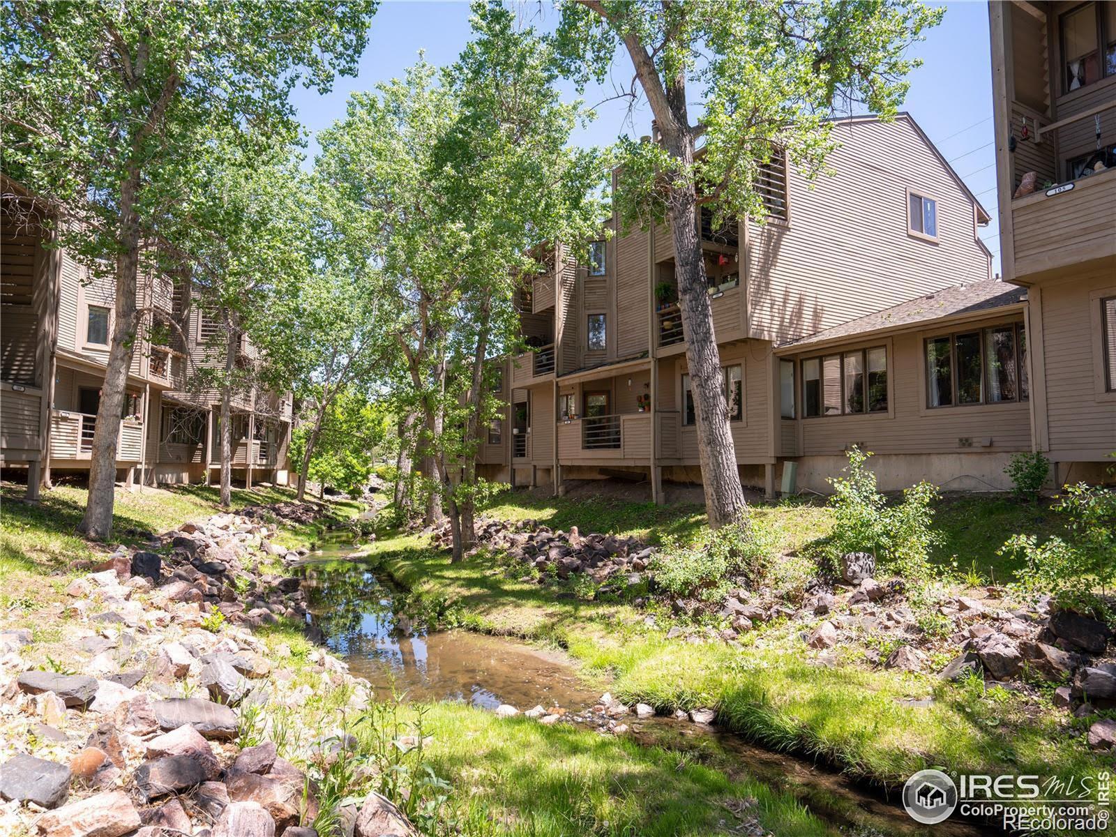Condominium buildings beside a rocky creek beneath mature leafy trees.