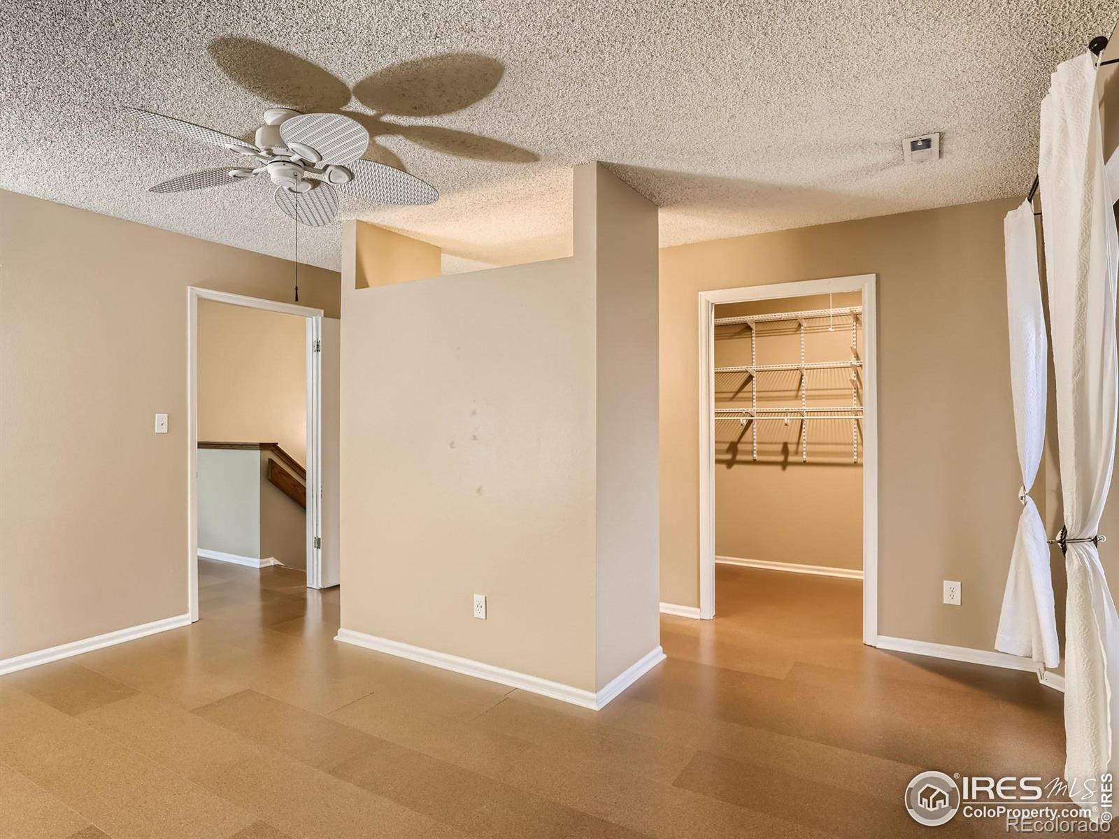 Empty bedroom with tan flooring, ceiling fan, white curtains, and an open closet with wire shelving.