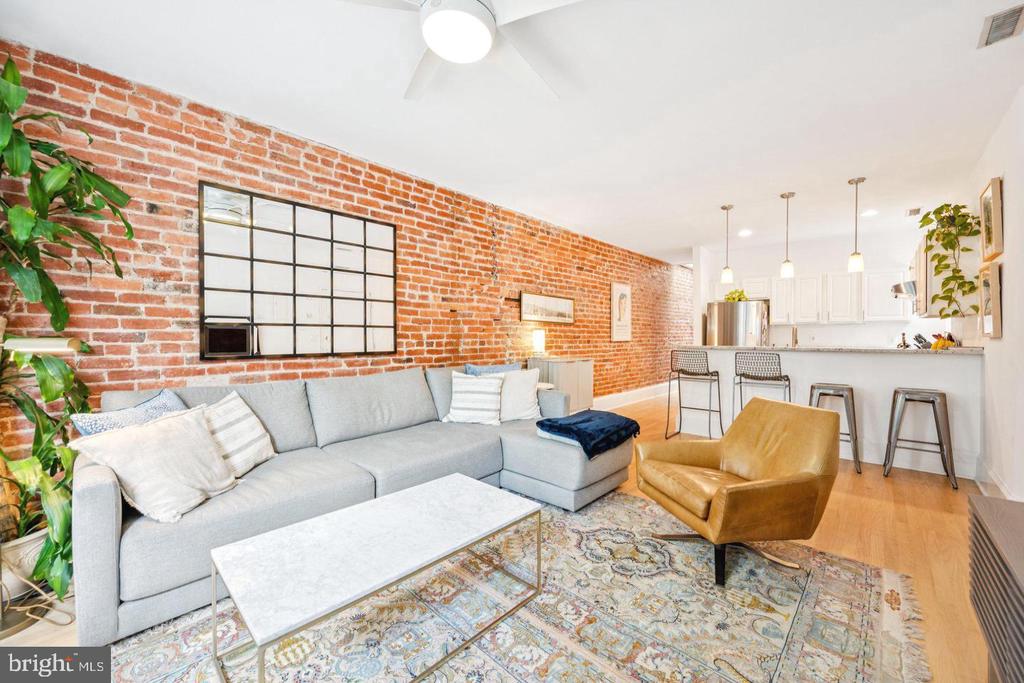 Open-plan living room with exposed brick wall, gray sectional, tan leather chair, and kitchen breakfast bar.