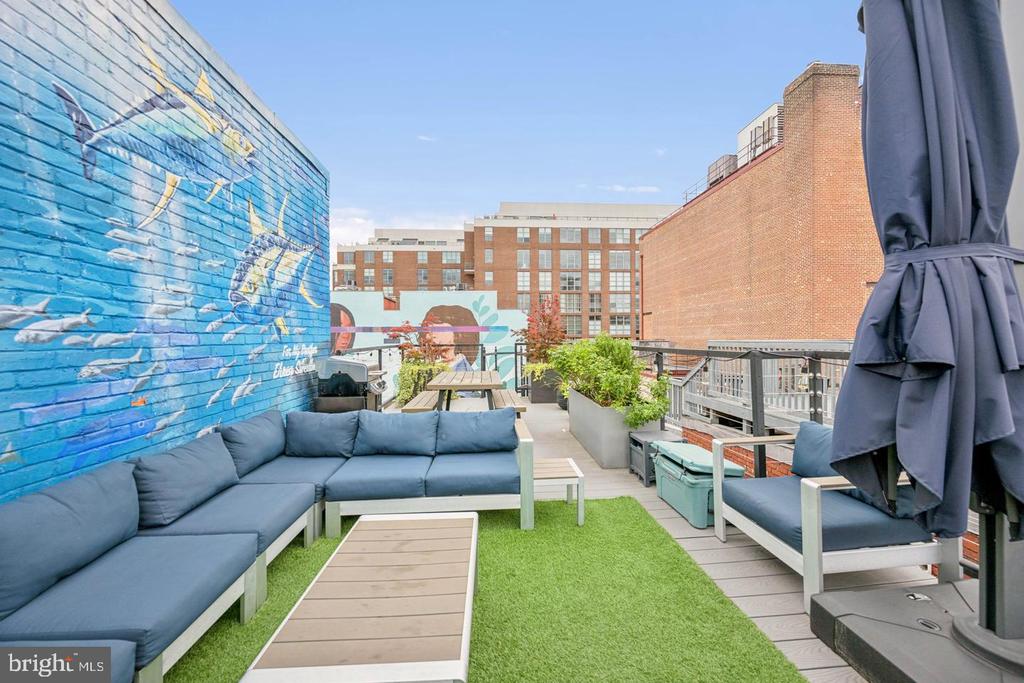 Rooftop terrace with a blue mural wall, sectional seating, artificial turf, and nearby city buildings.