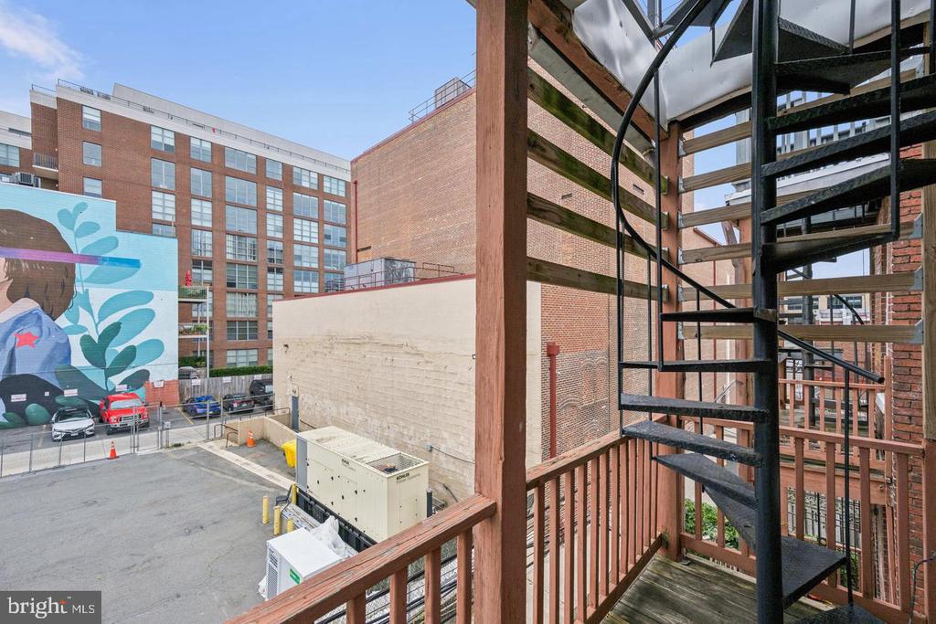 Balcony with a black spiral staircase overlooking brick buildings and a colorful mural.