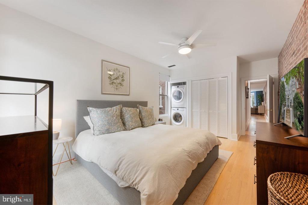 Bedroom with gray upholstered bed, stacked washer and dryer, louvered closet doors, and exposed brick wall.