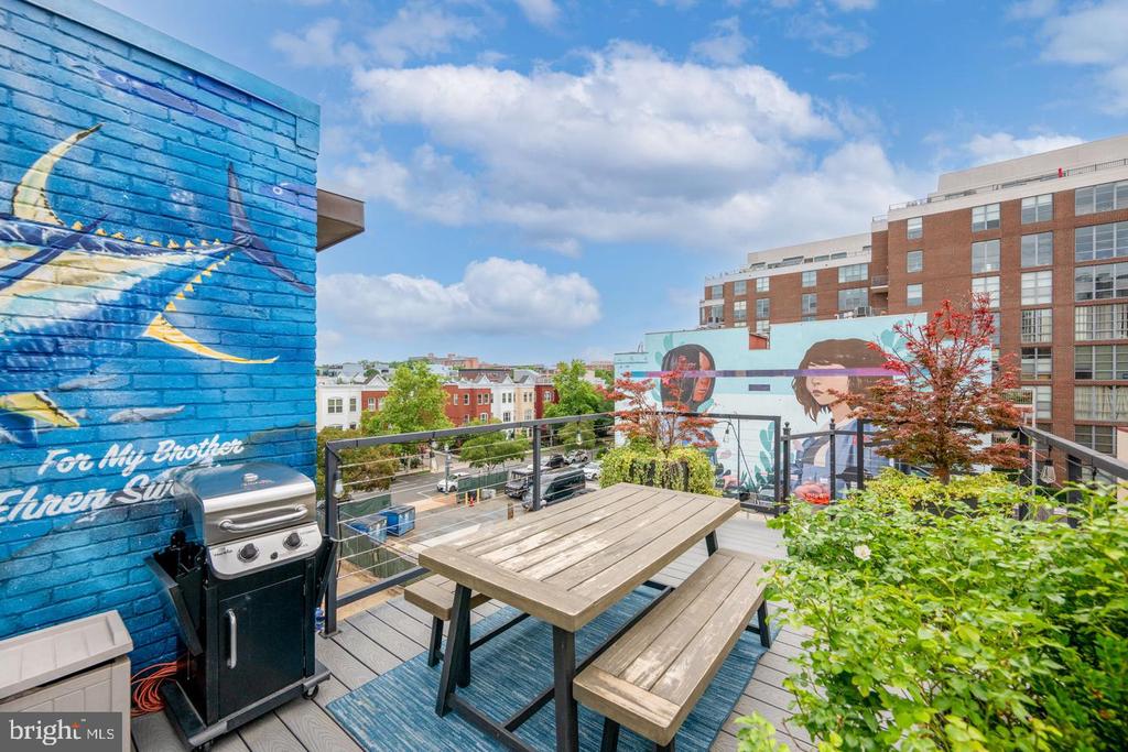 Rooftop deck with a wooden picnic table, grill, colorful murals, and views of nearby buildings.