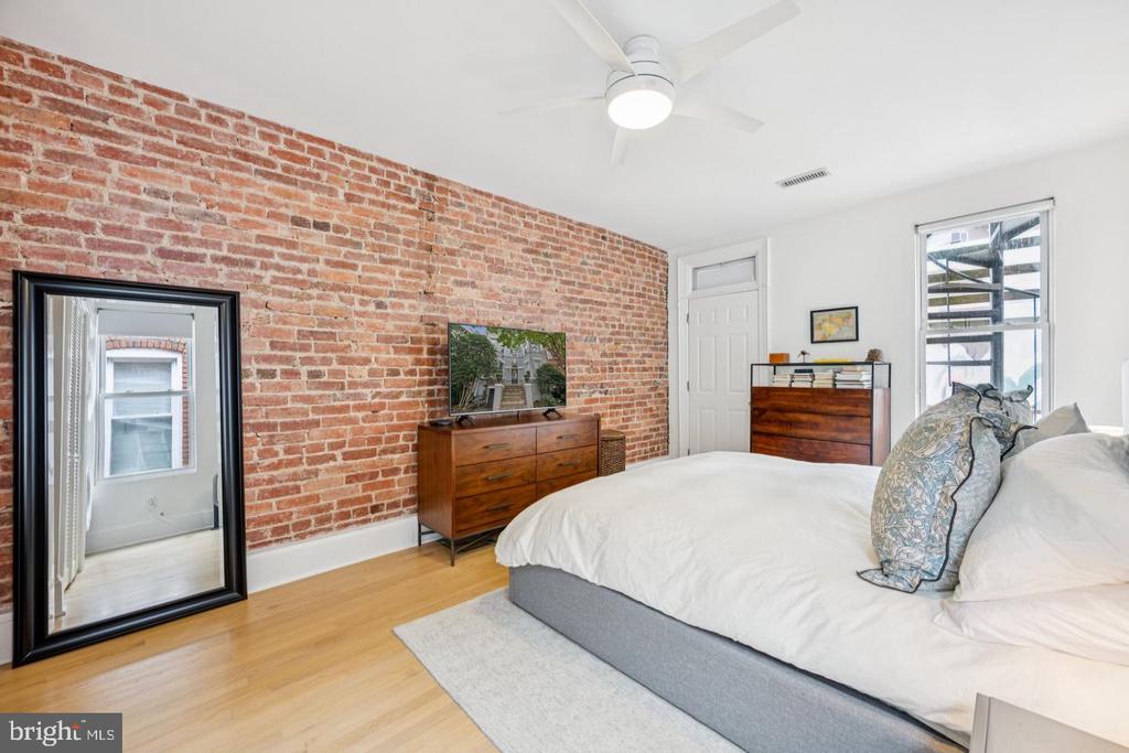 Bedroom with an exposed brick wall, white bed, and large black-framed floor mirror.