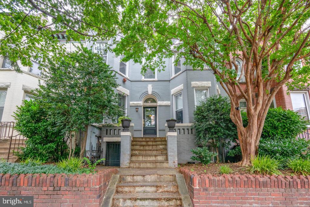 Gray townhouse with an arched entrance, stone steps, brick walls, and leafy trees.