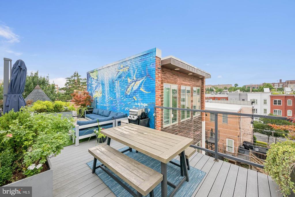 Rooftop terrace with a wooden picnic table, blue sofa, and colorful mural overlooking neighboring buildings.