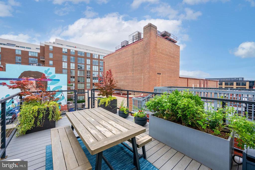 Rooftop terrace with a wooden dining table, planters, and surrounding city buildings.