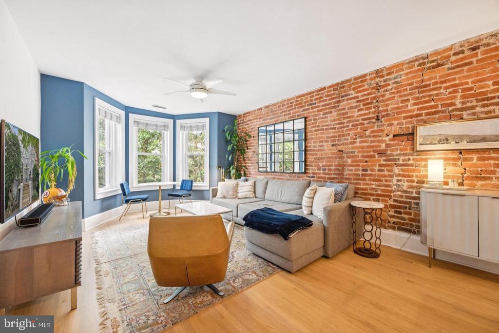 Living room with exposed brick wall, blue window alcove, gray sectional, and hardwood floors.