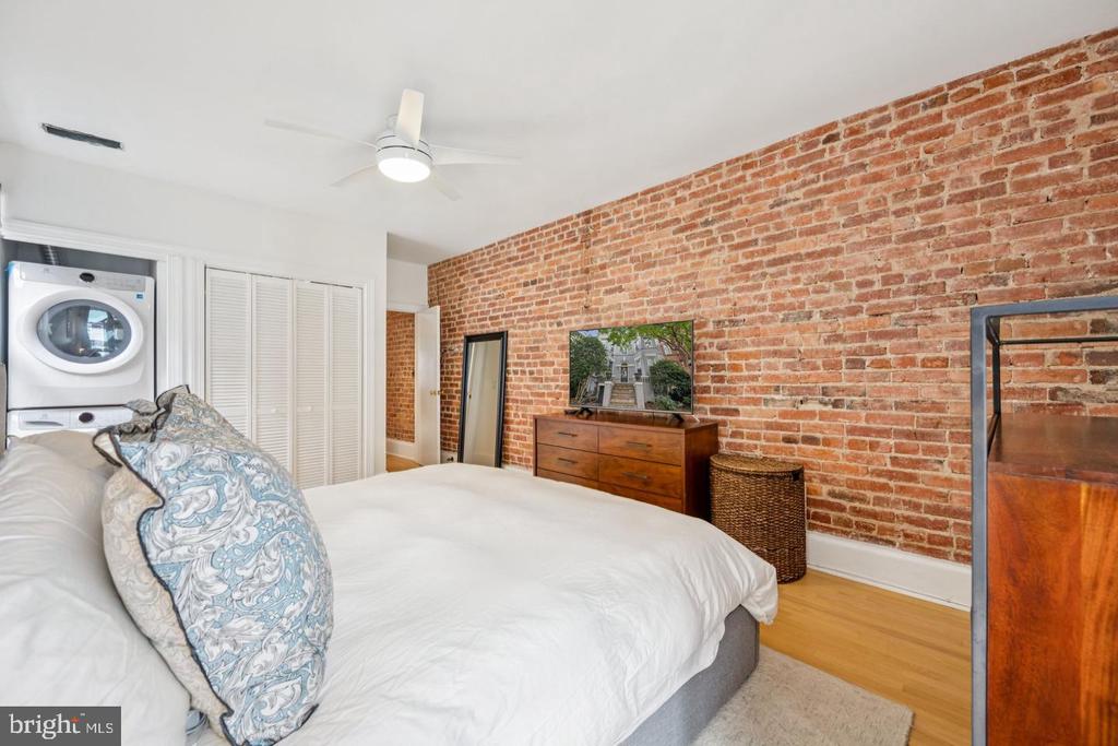 Bedroom with exposed brick wall, white bed, and stacked washer and dryer.