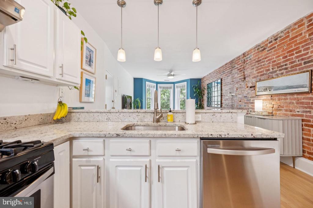 Kitchen with exposed brick wall, white cabinets, stone counters, and stainless-steel dishwasher.