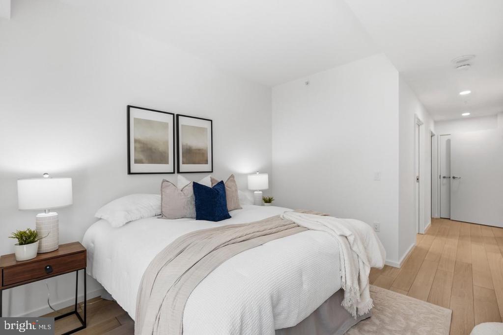 Staged bedroom with a white bed, navy accent pillow, framed prints, and a hallway with light wood floors.