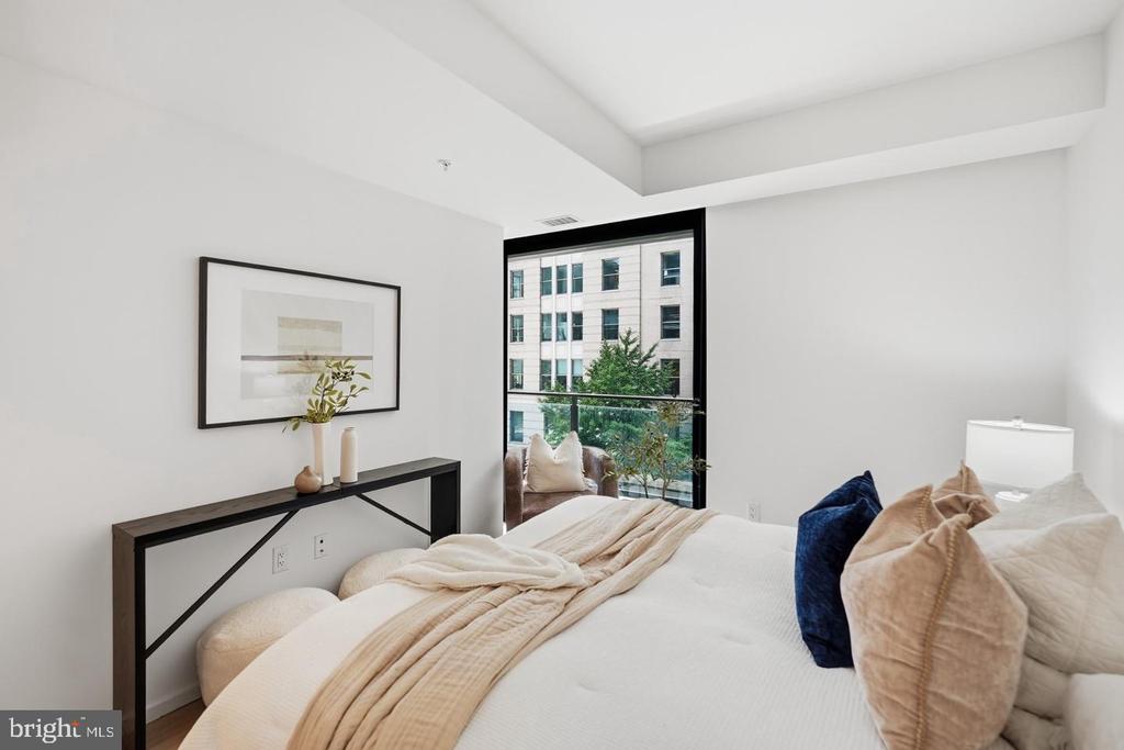 Staged bedroom with a white bed, black console table, and large window overlooking a neighboring building.