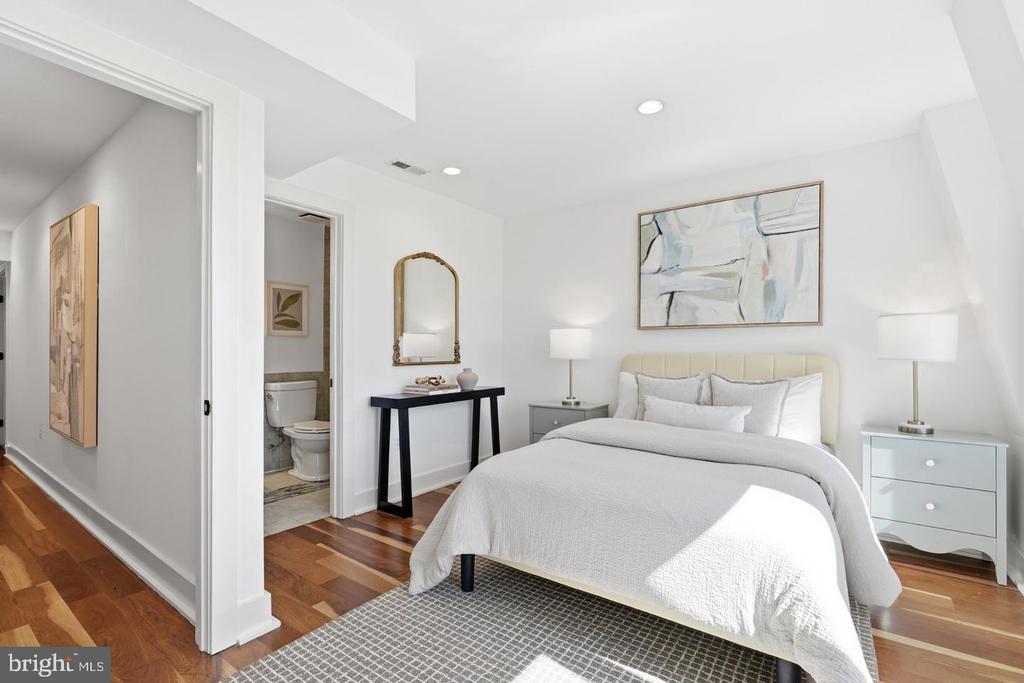 Staged bedroom with a white bed, hardwood floors, and an open doorway to the bathroom.