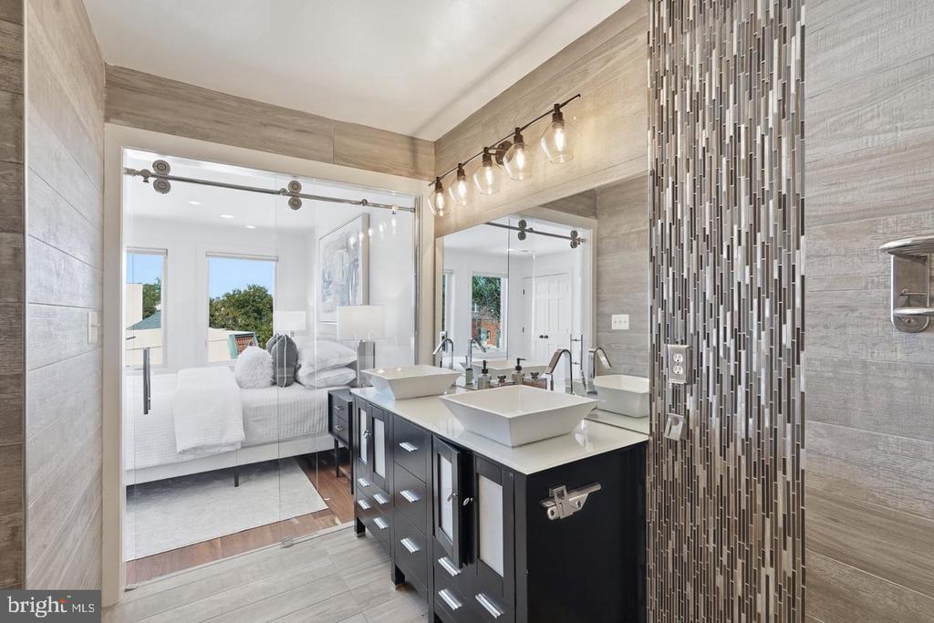 Modern bathroom with a dark double vanity, vessel sinks, and a glass partition opening to a bedroom.