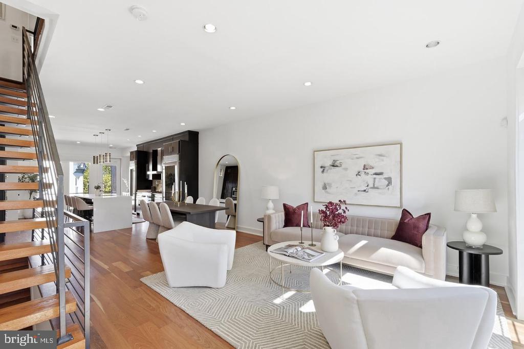 Bright staged living room with hardwood floors, white seating, and an open kitchen-dining area.