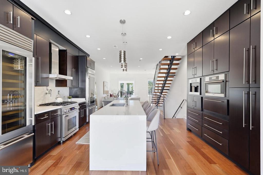 Modern kitchen with dark cabinetry, stainless appliances, a white island, and an open-riser staircase.