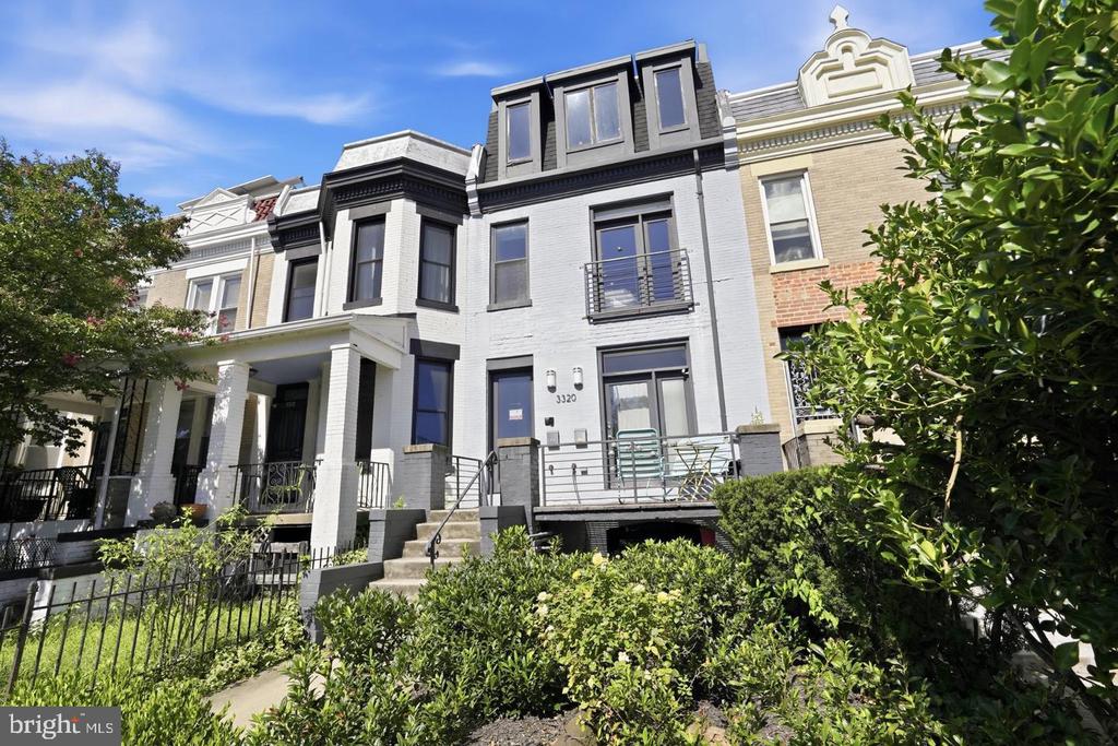 White-painted row house with dark-framed windows, front stoop, and leafy landscaping.