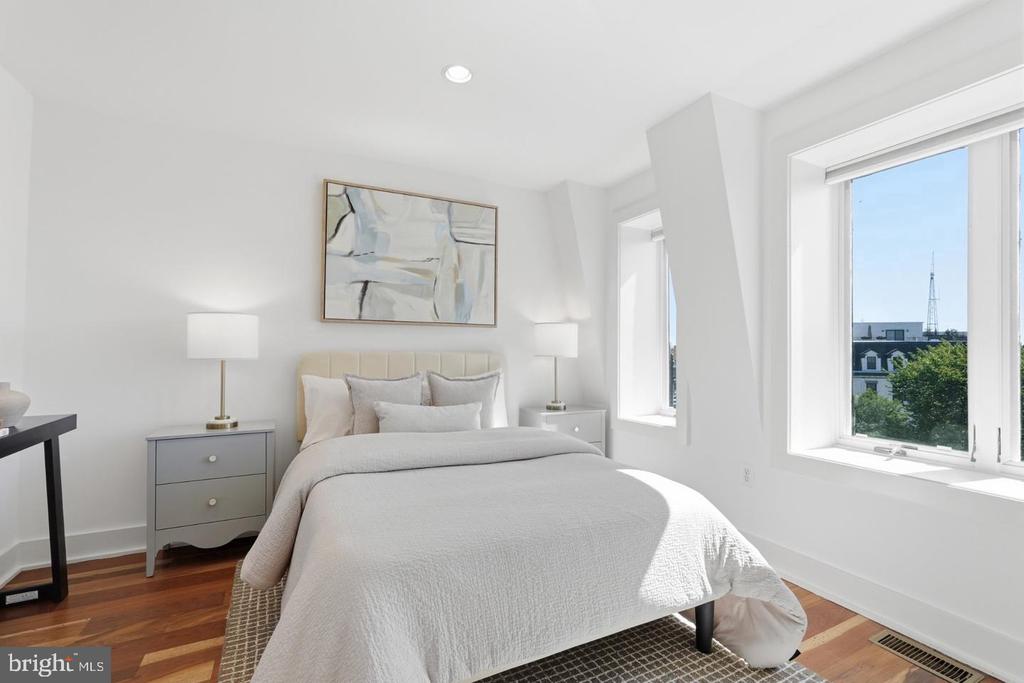 Bedroom with a white bed, hardwood floors, and tall windows overlooking rooftops and trees.