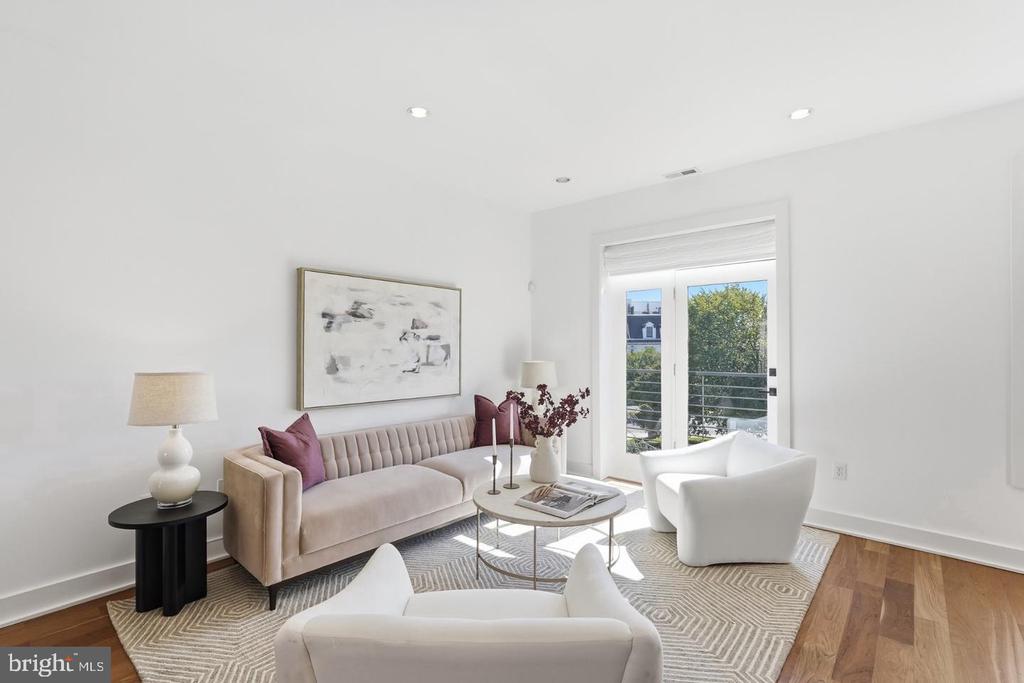 Staged living room with a beige tufted sofa, white chairs, and glass doors to a balcony.