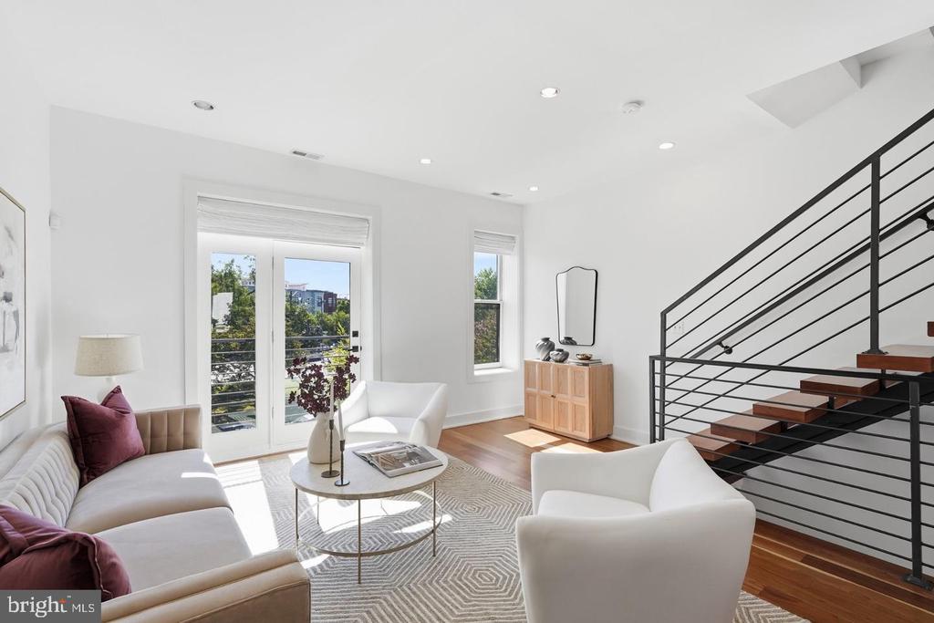Living room with white seating, balcony doors, hardwood floors, and an open staircase.