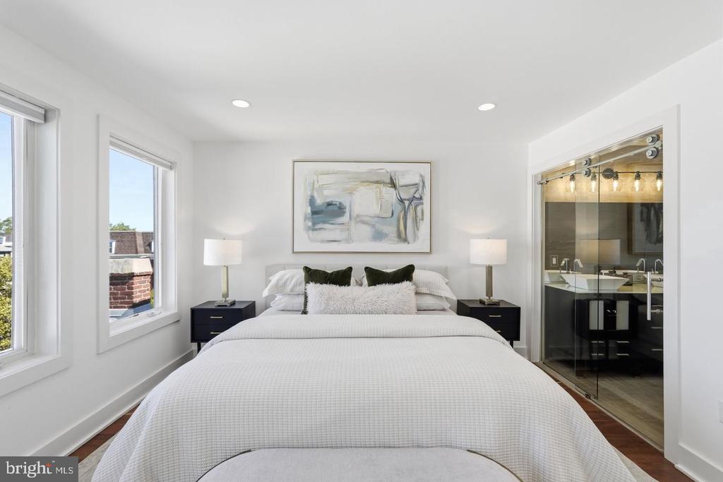 Staged bedroom with a white bed, two windows, and a glass-walled ensuite bathroom.