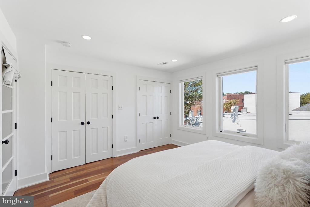 Bright bedroom with hardwood floors, a large bed, and tall windows overlooking nearby rooftops.