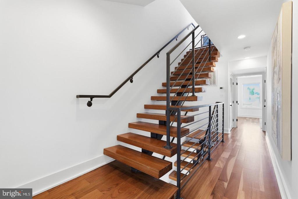 Open-riser wood staircase with dark metal railings beside a bright hardwood hallway.
