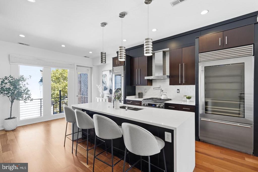 Modern kitchen with a long white island, four gray stools, dark cabinets, and stainless steel appliances.