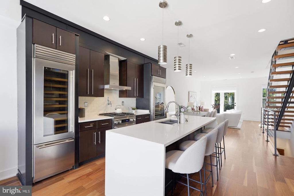 Modern kitchen with a large white island, dark cabinetry, stainless appliances, and an open staircase.