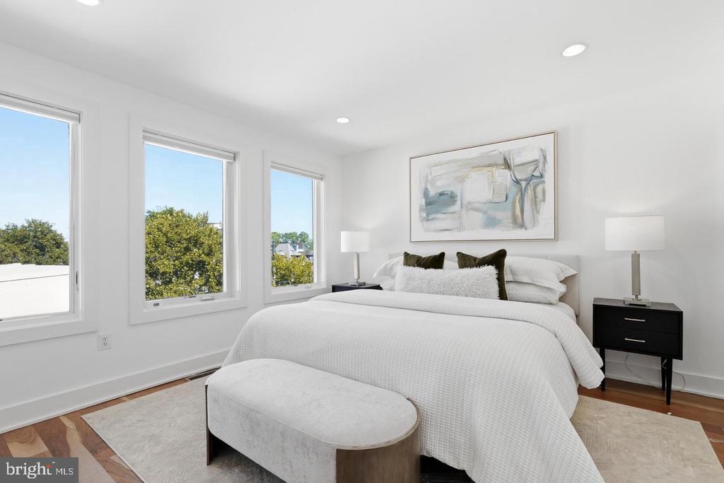 Staged bedroom with a white bed, upholstered bench, abstract artwork, and three tall windows overlooking trees.