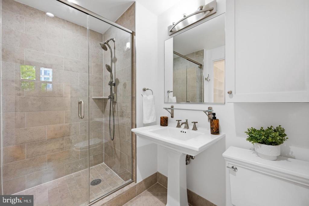 Bathroom with a beige tiled glass shower, pedestal sink, mirrored cabinet, and potted plant.