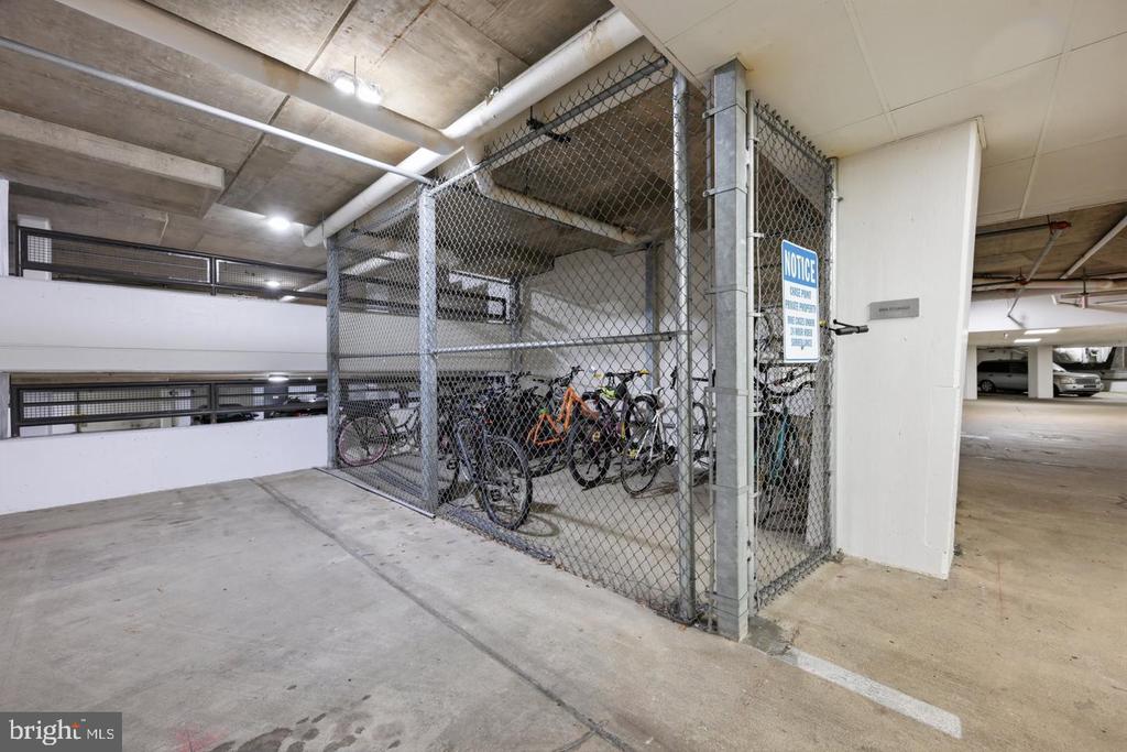 Bicycle storage cage in a concrete parking garage, with several bikes behind chain-link fencing.