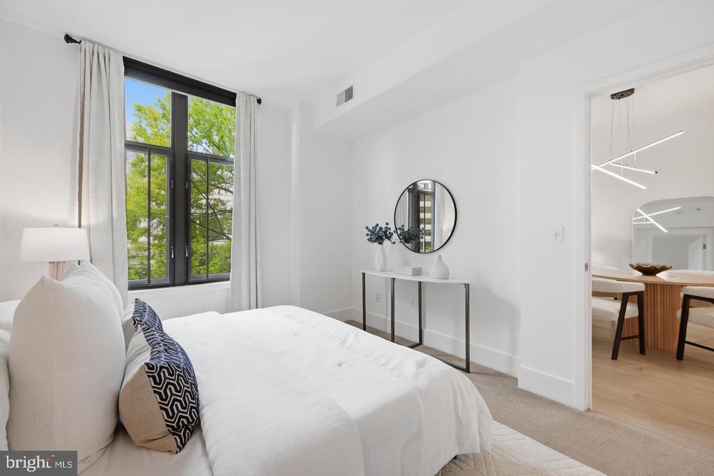 Staged bedroom with a white bed, tall black-framed window, and adjoining dining area.