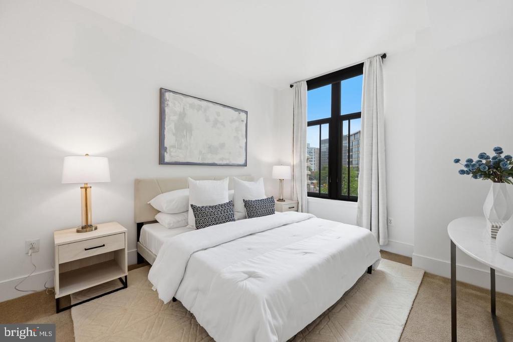 Staged bedroom with a white bed, patterned pillows, and a large window overlooking nearby buildings.