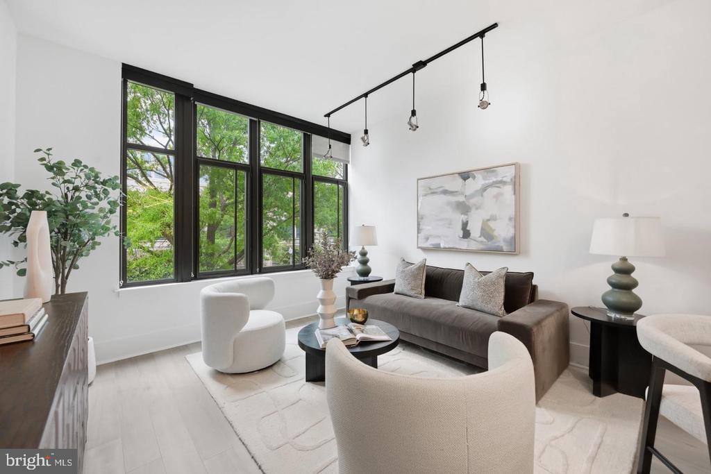 Staged living room with black-framed windows, leafy views, a gray sofa, and white chairs.