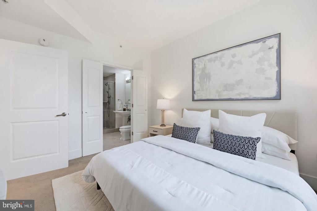 Staged bedroom with a white bed, patterned pillows, and abstract artwork; en-suite bathroom visible through doorway.