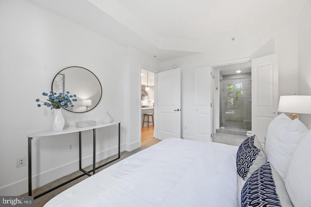 Bedroom with a white bed, round mirror, and open doors to an en-suite bathroom and kitchen.