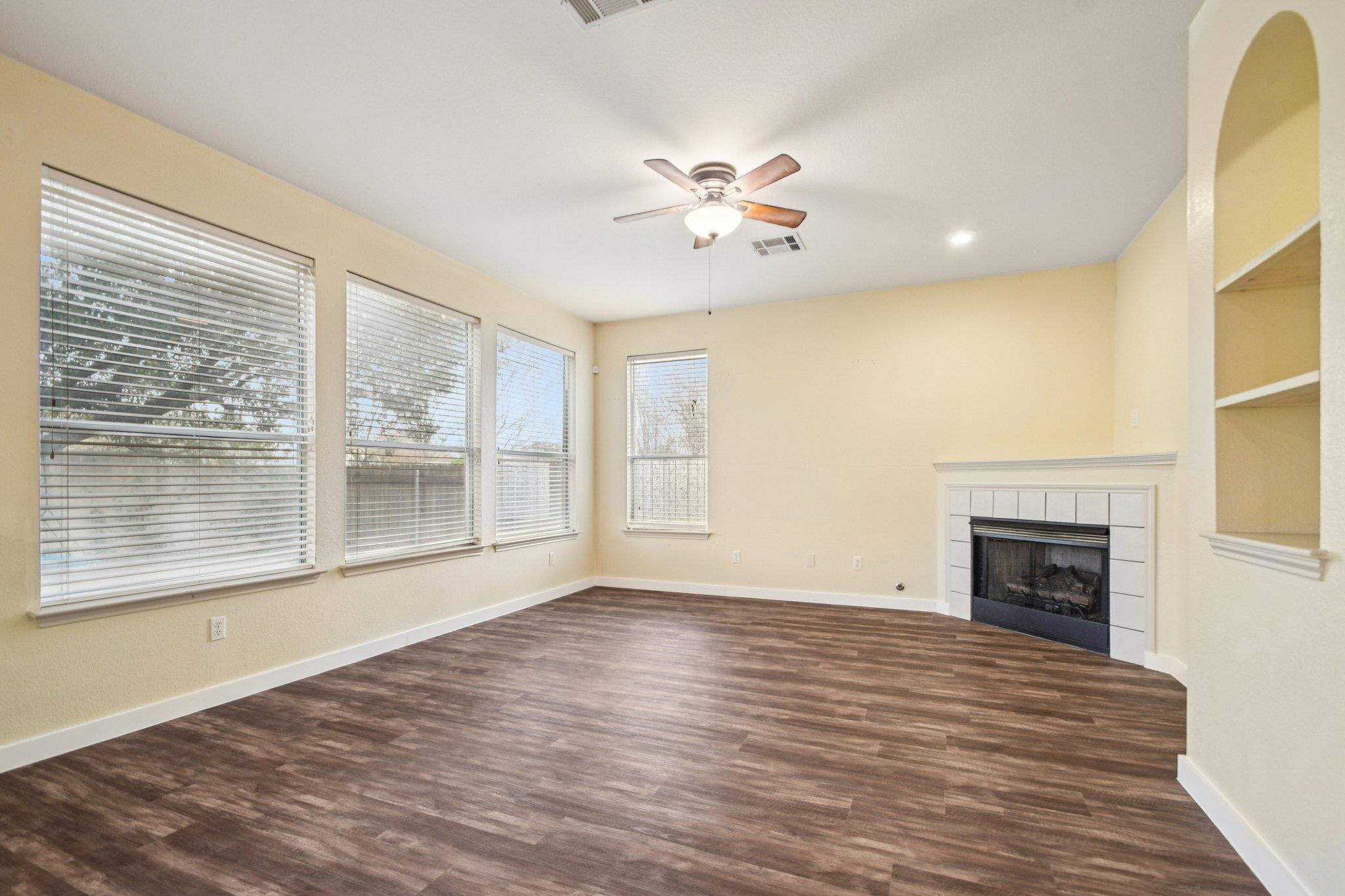 Empty living room with dark wood-look floors, multiple windows, a ceiling fan, and a tiled fireplace.