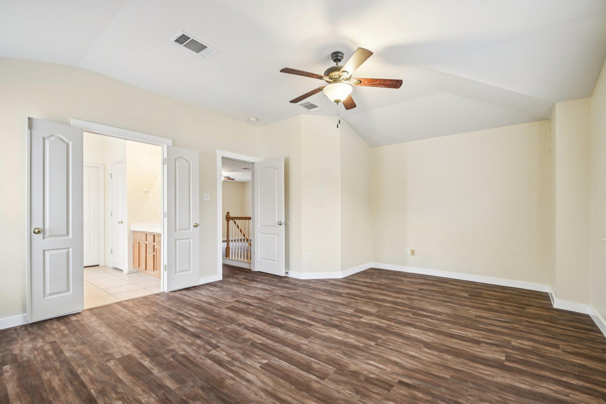 Empty bedroom with wood-look flooring, a ceiling fan, and open doors to a bathroom and hallway.
