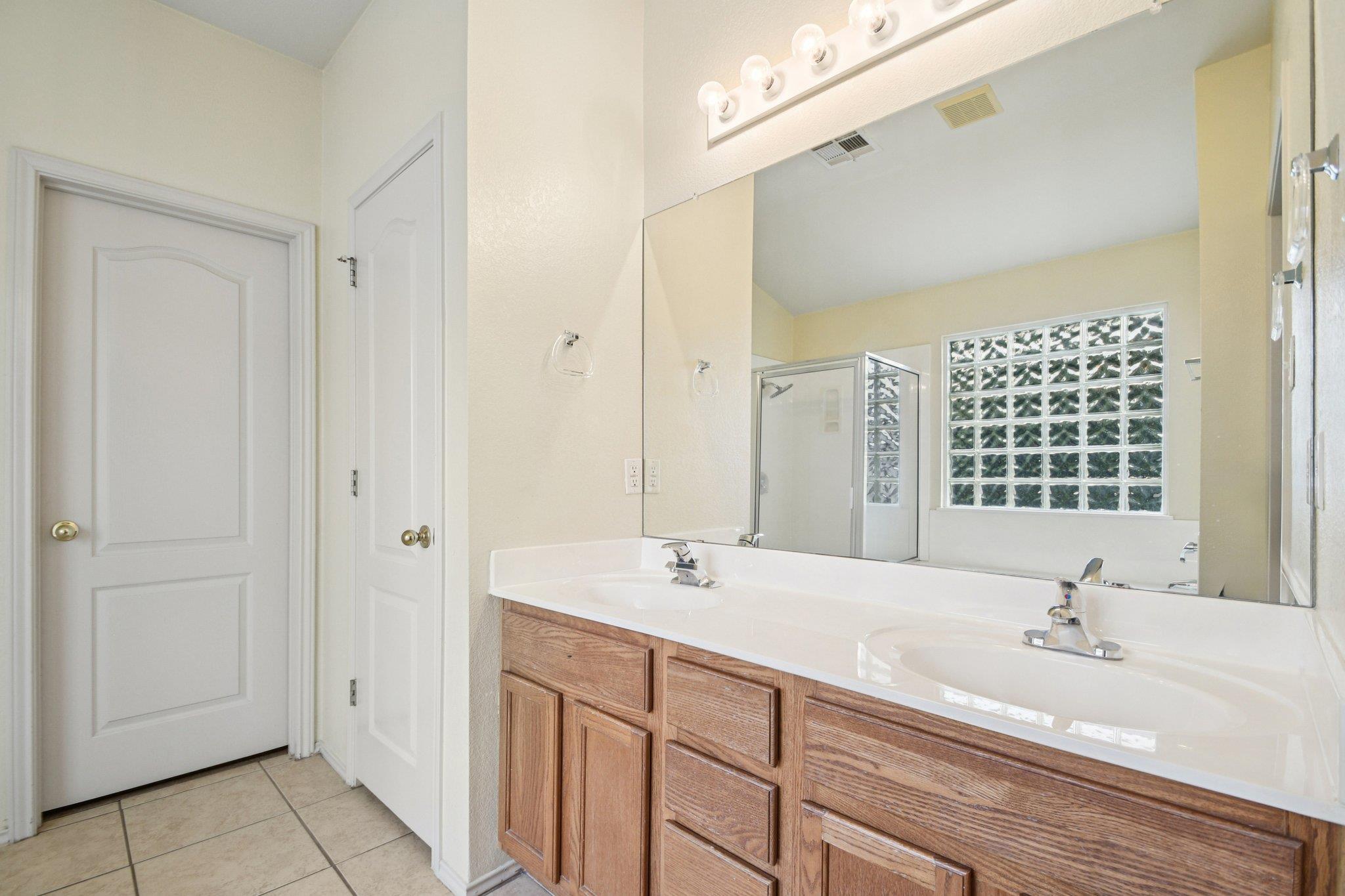 Bathroom with a double-sink wood vanity, large mirror, glass-block window, and glass-enclosed shower.