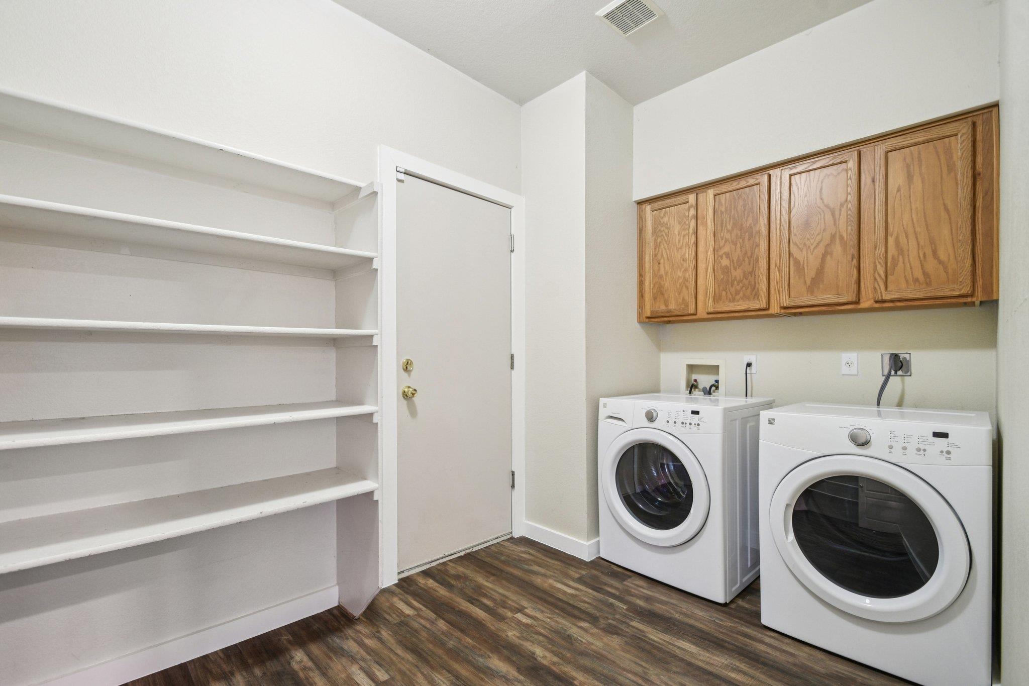 Laundry room with front-loading washer and dryer beside built-in shelving and wood cabinets.