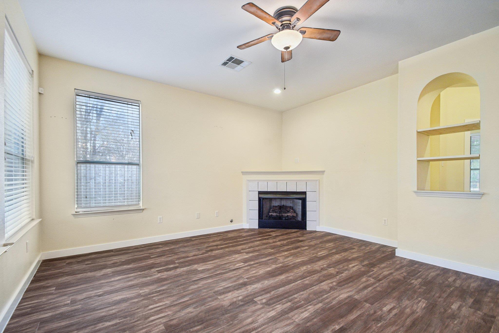 Empty living room with a tiled fireplace, arched built-in shelves, and dark wood-look flooring.