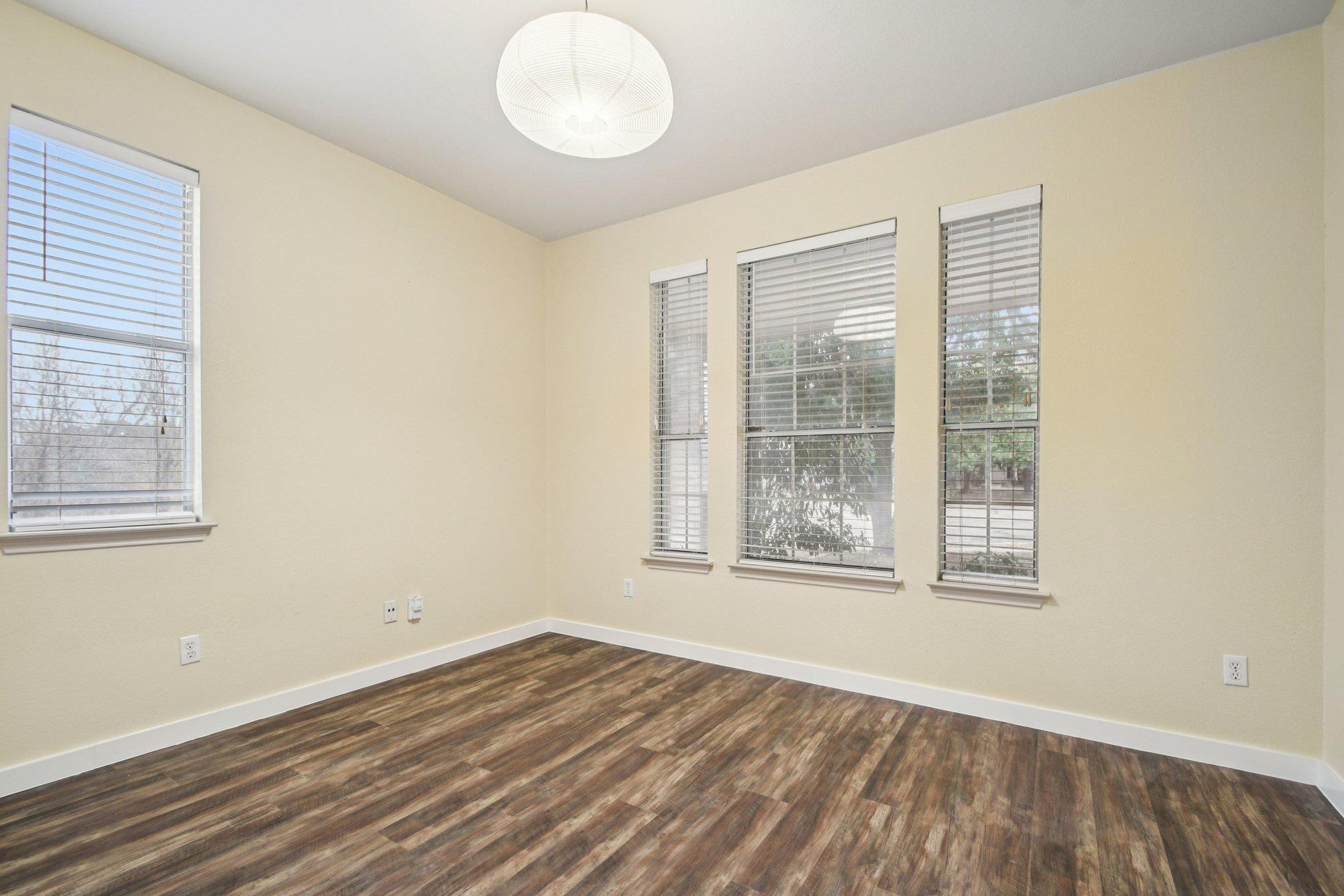 Vacant room with dark wood-look flooring, tall blinded windows, and a white pendant light.