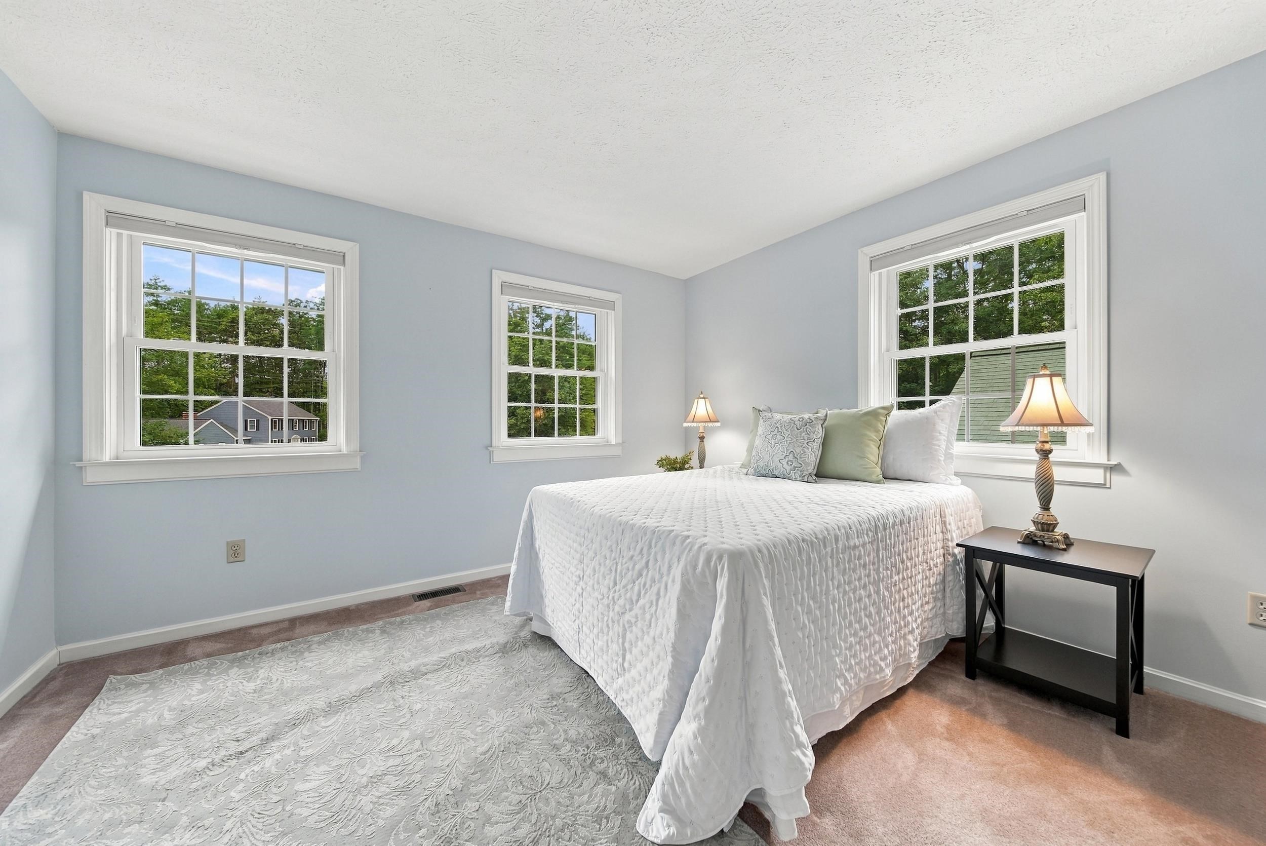 Bright bedroom with a white quilted bed and three large windows overlooking trees.