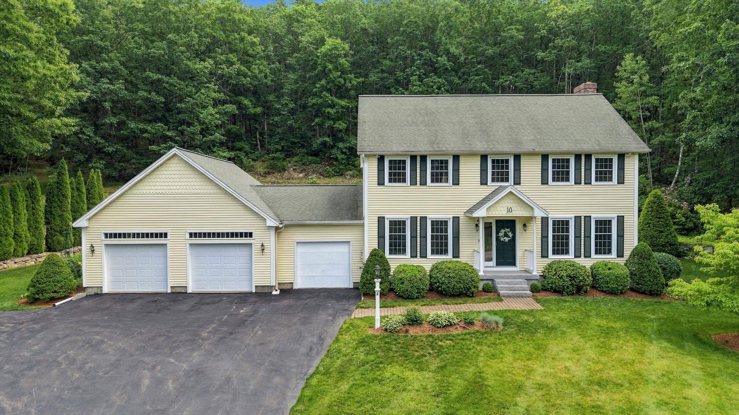 Pale yellow two-story house with three garage doors, a landscaped lawn, and dense woods behind it.